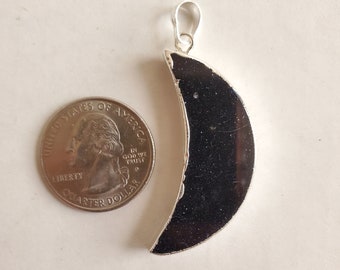May include: A crescent moon pendant with a silver-colored frame and a dark, possibly black, center. The pendant is next to a U.S. quarter for size comparison. The pendant has a loop at the top for a chain.