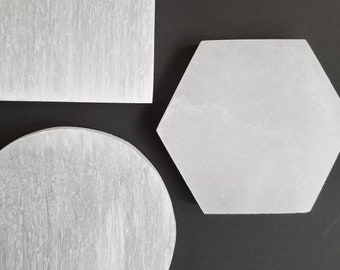 May include: Three white selenite crystal shapes: a square, a hexagon, and a circle. A coin is next to the shapes for size comparison. The shapes are smooth and have a natural, translucent appearance.