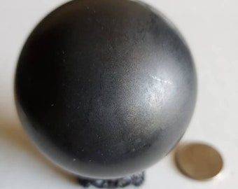 Puede incluir: Una esfera gris oscuro con una superficie ligeramente texturizada. Un pequeño objeto redondo de color dorado está al lado de la esfera. La esfera parece ser de metal. La esfera tiene aproximadamente 7,6 cm de diámetro.