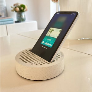 Peut inclure: Un support de téléphone en bois blanc avec un motif en grille et le mot "PhoneRack" imprimé sur le côté. Un smartphone est placé dans le support, affichant une application de streaming musical avec une scène de plage.