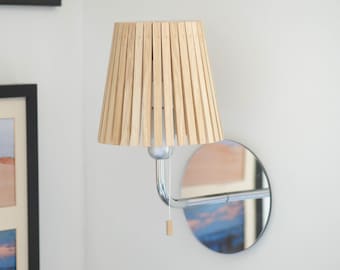 May include: A wall-mounted light fixture with a wooden slat shade and a chrome arm. The shade is made of natural wood slats and has a round shape. The arm is attached to a round mirror.
