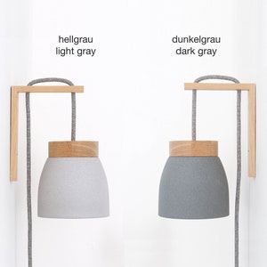 Könnte beinhalten: Zwei Wandleuchten mit Holzsockeln und grauen Stoffkabeln. Die Lampenschirme sind aus grauem, strukturiertem Material gefertigt. Der Text "hellgrau light gray" und "dunkelgrau dark gray" ist über jeder Leuchte angebracht.