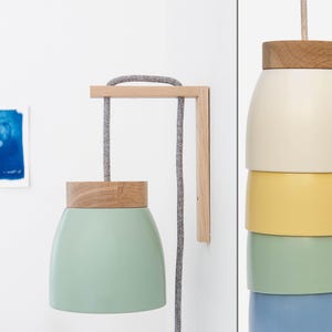 Könnte beinhalten: Eine Wandleuchte mit einem Holzrahmen und einem mehrschichtigen Schirm in Grün, Gelb, Blau und Weiß. Das Kabel ist grau und der Schirm ist aus Metall.