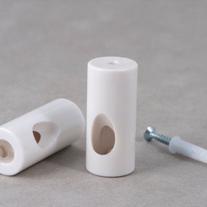 Puede incluir: Dos tacos de pared de plástico blanco con un agujero para tornillo en la parte superior y un gran agujero ovalado en el lateral. También se muestra un taco de tornillo de plástico blanco con un tornillo plateado.