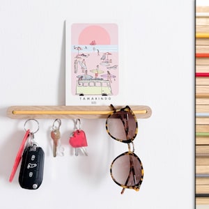 May include: A light-colored wooden wall-mounted organizer featuring a horizontal yellow cord. A rectangular print titled TAMARINDO depicting a beach scene rests on the shelf. Hanging from the cord are three sets of keys and tortoise-shell sunglasses.
