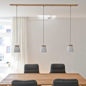 May include: Three white pendant lights with wooden tops hang from a wooden rail in a dining room. The lights are over a wooden table with four grey chairs.