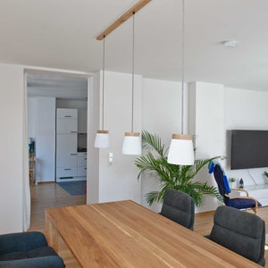 May include: A wooden dining table with a light wood finish and three pendant lights hanging above it. The table is surrounded by four chairs with dark gray fabric upholstery. A potted plant is in the background.