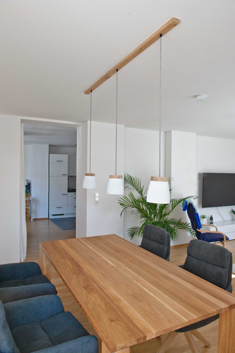 May include: A wooden dining table with a light wood finish and three pendant lights hanging above it. The table is surrounded by four chairs with dark gray fabric upholstery. A potted plant is in the background.