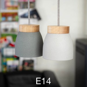 Op de afbeelding: Twee hanglampen met houten toppen en grijze en witte stoffen lampenkappen. De lampen hangen aan snoeren en de tekst "E14" is zichtbaar onder de lampen.