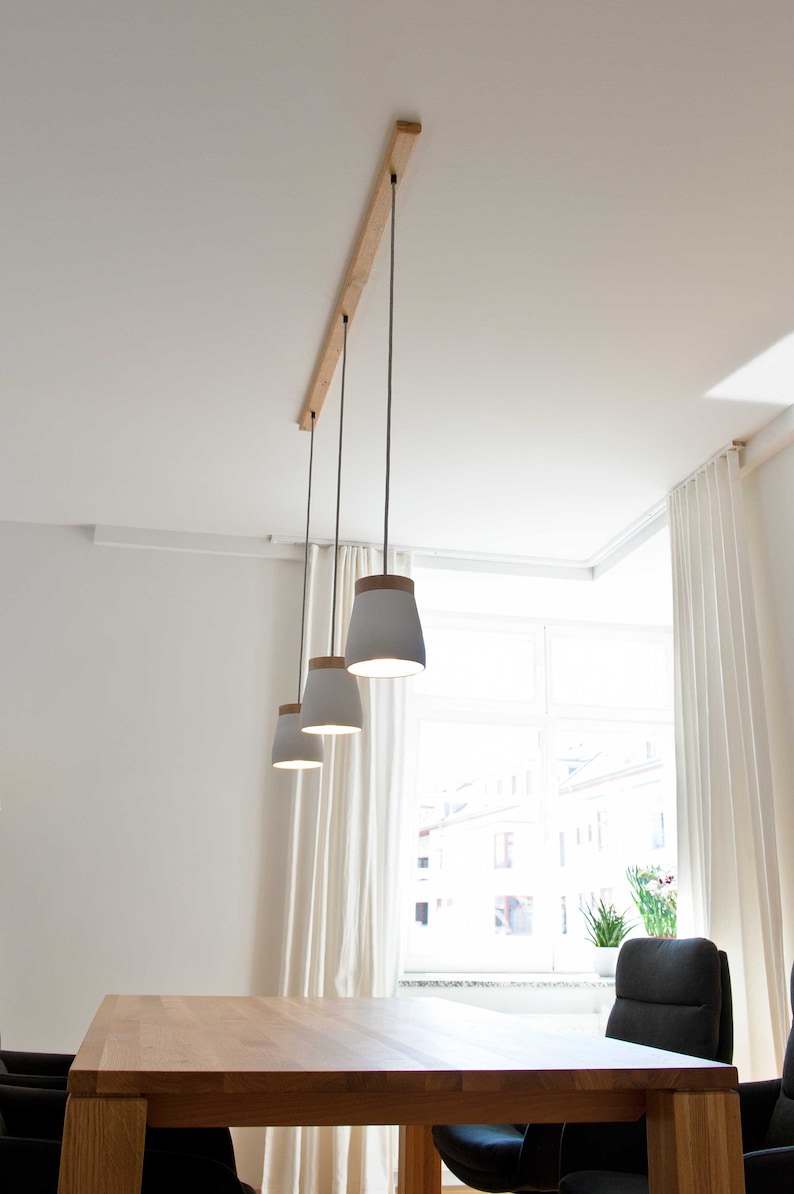 May include: Three white pendant lights with wooden tops hang from a wooden rail on a white ceiling. The lights are over a wooden table with black chairs.