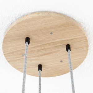 Peut inclure: Plafonnier en bois clair avec trois serre-câbles noirs et des câbles électriques recouverts de tissu gris. Le support de plafond circulaire est fixé contre un plafond blanc. Les câbles suggèrent un luminaire suspendu.