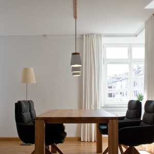 May include: A wooden dining table with four black chairs and a white pendant light fixture with three shades hanging from the ceiling. The table is set in a room with white walls and a window with white curtains.