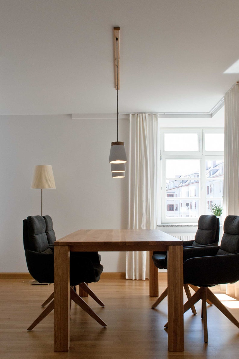 May include: A wooden dining table with four black chairs and a white pendant light fixture with three shades hanging from the ceiling. The table is set in a room with white walls and a window with white curtains.