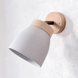 以下が含まれることがあります： ライトグレーの円錐形シェードと天然木ベースを備えたモダンなウォールライト。白い壁に取り付けられ、シェードと木製ベースを繋ぐダークメタルアームが特徴です。ミニマルで現代的なデザインです。