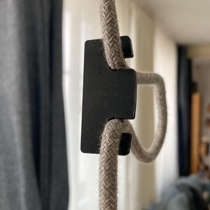 May include: A close-up of a black cord clamp holding a beige braided cord. The cord is looped through the clamp and is hanging from the ceiling.