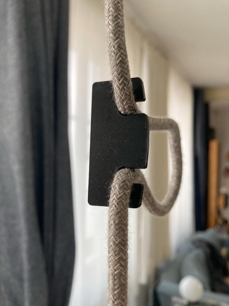 May include: A close-up of a black cord clamp holding a beige braided cord. The cord is looped through the clamp and is hanging from the ceiling.