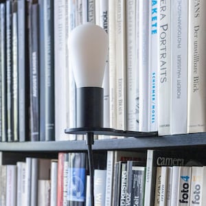 May include: A modern black desk lamp with a white bulb, positioned on a shelf filled with books. The lamp has a sleek design with a black base and a curved arm. The bulb is a soft white color.