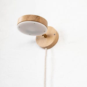 May include: A wooden wall-mounted light fixture with a white diffuser and a cord hanging down. The light is made of natural wood and has a simple, modern design.