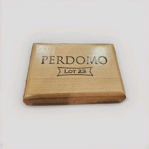 May include: A rectangular wooden cigar box with the text "PERDOMO LOT 23" printed on the top. The box is light brown with rounded edges and the text "CONNECTICUT" and "ROBUSTO" printed on the side.