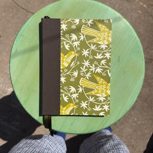 May include: A rectangular notebook featuring a dark brown spine and a cover with a repeating pattern of yellow birds and white botanical elements on an olive green background. A dark green ribbon bookmark extends from the bottom of the spine.