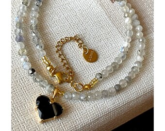 Op de afbeelding: Een dubbele ketting van grijze edelsteen kralen met een gouden ketting en een zwarte hart hanger. De hanger heeft een gouden rand. Het label op de ketting luidt "925 STERLING".