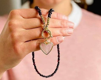 Op de afbeelding: Een gouden ketting met een hartvormige hanger. De hanger is gemaakt van lichtroze materiaal en heeft een gouden rand. De ketting is gemaakt van kleine zwarte kralen en gouden kralen.