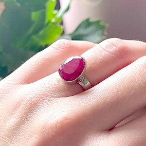 Puede incluir: Un anillo de plata con una gran piedra preciosa de rubí de forma ovalada. El rubí es de color rojo intenso y tiene un acabado liso y pulido.