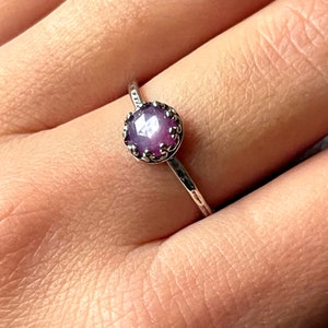 Peut inclure: Une bague en argent avec une pierre précieuse violette sertie dans une fine bélière détaillée. La bague est portée à un doigt.