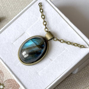 Könnte beinhalten: Ovale Anhänger Halskette mit einem Labradorit-Stein. Der Stein hat blau-grüne schillernde Farben, gefasst in einem bronzefarbenen Metallrahmen. Die Kette ist ebenfalls bronzefarben und liegt in einer weißen Schmuckschatulle.