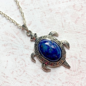 Può includere: Una collana con un ciondolo a forma di tartaruga in argento, decorata con una pietra di lapislazzuli blu incastonata in una cornice decorativa. Il ciondolo presenta un design di tartaruga dettagliato con un bordo a chiave greca.