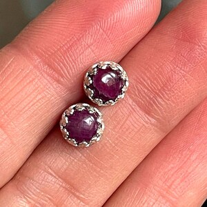 May include: A pair of silver stud earrings featuring deep red ruby gemstones. Each ruby is set in a silver bezel with a decorative border.