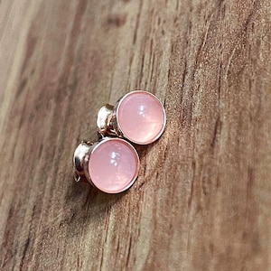 Op de afbeelding: Een paar rozenkwarts oorknopjes. Elke oorbel heeft een ronde, gepolijste roze edelsteen, gezet in een zilverkleurige metalen bezel. De oorbellen worden gepresenteerd op een houten oppervlak.