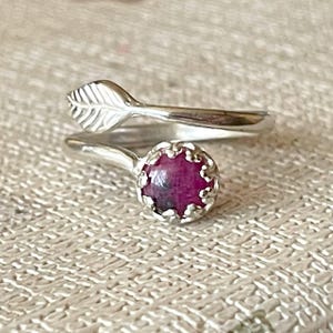 Puede incluir: Un anillo plateado con un diseño de hoja y una piedra preciosa redonda de color rosa intenso. La banda del anillo se envuelve alrededor del dedo, con el detalle de la hoja en un extremo y la piedra preciosa engastada en un bisel decorativo en el otro. La gema mide unos 0,8 cm de diámetro.