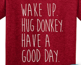以下が含まれることがあります： 「WAKE UP. HUG DONKEY. HAVE A GOOD DAY.」と白い文字で書かれたマルーン色のTシャツ。右下には馬のグラフィックが入った円形のロゴがあります。このシャツはクルーネックで半袖です。