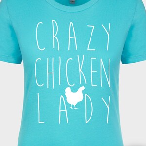 Peut inclure: Un t-shirt turquoise avec l'inscription blanche "CRAZY CHICKEN LADY" et un dessin de poulet blanc. Le t-shirt a un col rond et des manches courtes. Un logo circulaire avec un cheval est dans le coin inf&eacute;rieur droit.
