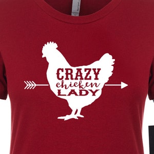 May include: A red t-shirt featuring a white graphic of a chicken with the text "CRAZY chicken LADY" in a serif font. An arrow points from left to right through the chicken. A black tag with a horse logo hangs from the shirt.