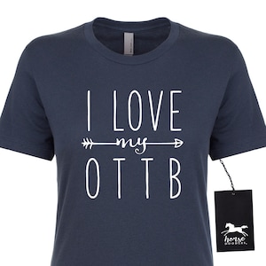 May include: Navy blue t-shirt with the white text "I LOVE my OTTB" and an arrow design. The shirt has a tag that says "horse doodles." The shirt is a short-sleeved crew neck.