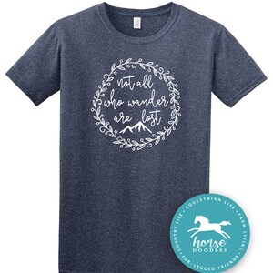 以下が含まれることがあります： 紺色のTシャツ。白い花のリースの中に「not all who wander are lost」という文字が書かれています。その下に山のグラフィックがあります。右下には、白い馬のグラフィックと「horse doodles」という文字が入ったターコイズ色の丸いステッカーがあります。