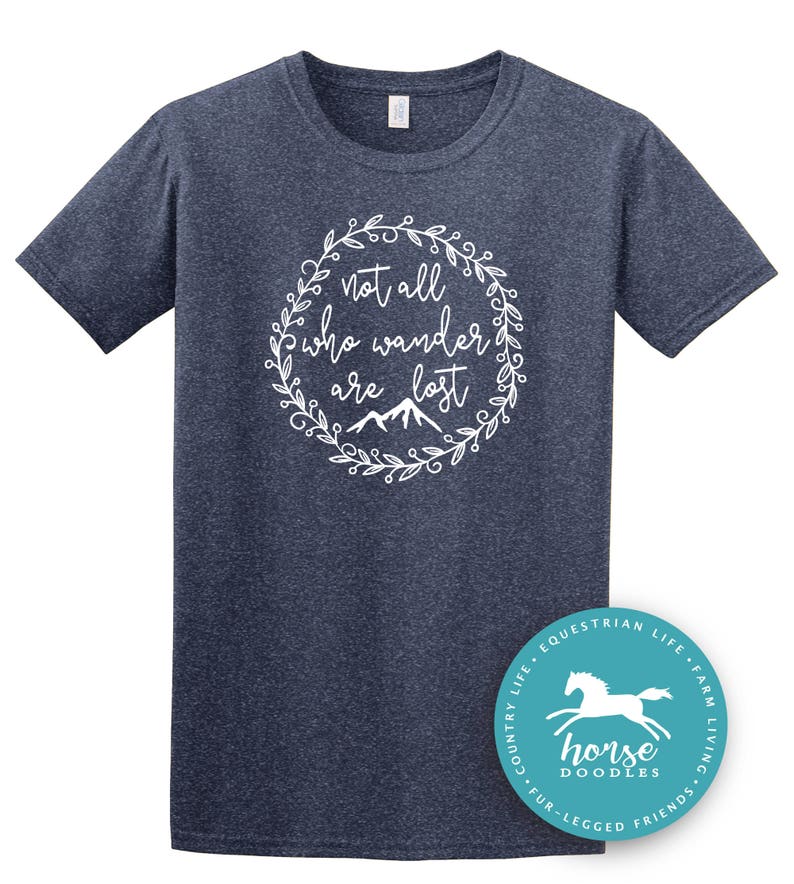 以下が含まれることがあります： 紺色のTシャツ。白い花のリースの中に「not all who wander are lost」という文字が書かれています。その下に山のグラフィックがあります。右下には、白い馬のグラフィックと「horse doodles」という文字が入ったターコイズ色の丸いステッカーがあります。
