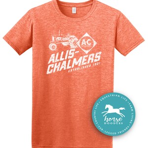 May include: Orange short-sleeved t-shirt with a distressed white graphic featuring a tractor and the text "ALLIS-CHALMERS ESTABLISHED 1901". A circular logo with a horse is on the bottom right.