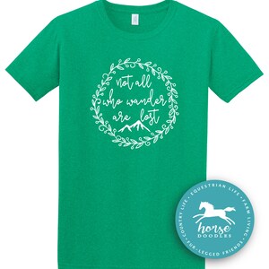 以下が含まれることがあります： 白い花のリースデザインで「not all who wander are lost」というテキストが入った緑色のTシャツ。シャツには、馬と「Horse Doodles」という言葉が入った円形のロゴも付いています。