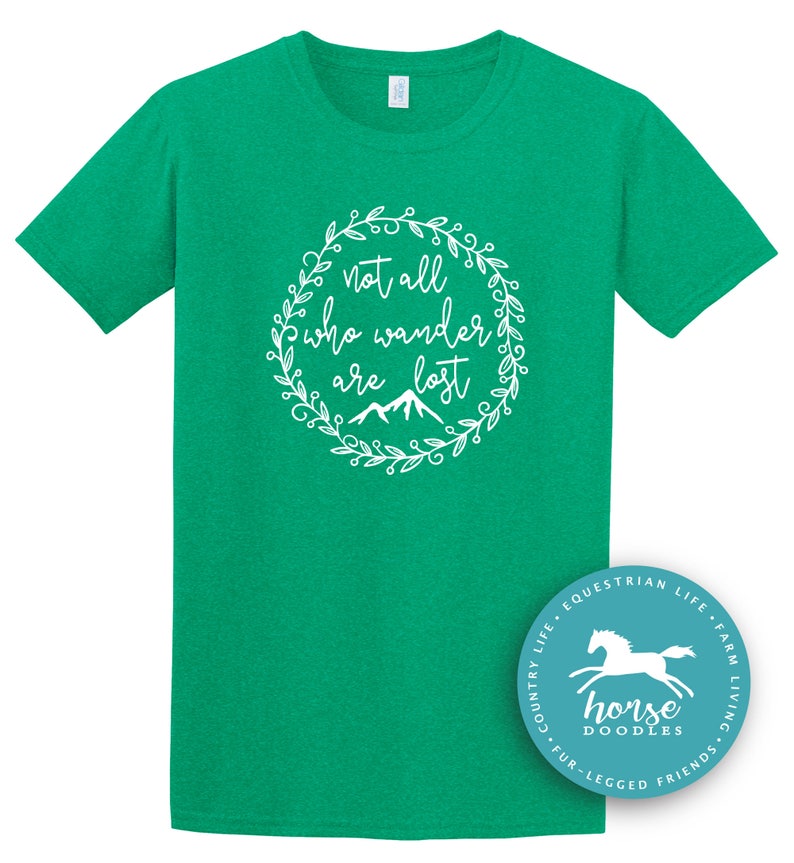 以下が含まれることがあります： 白い花のリースデザインで「not all who wander are lost」というテキストが入った緑色のTシャツ。シャツには、馬と「Horse Doodles」という言葉が入った円形のロゴも付いています。