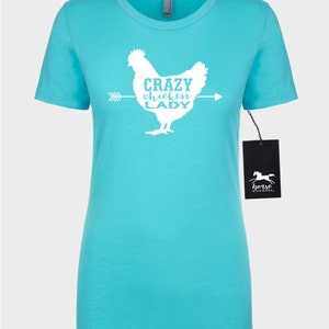 May include: A turquoise t-shirt featuring a white graphic of a chicken with the text "CRAZY CHICKEN LADY" and an arrow. The shirt has short sleeves and a crew neck. A black tag with a horse logo hangs on the side.