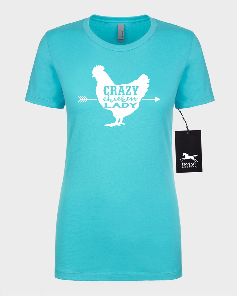May include: A turquoise t-shirt featuring a white graphic of a chicken with the text "CRAZY CHICKEN LADY" and an arrow. The shirt has short sleeves and a crew neck. A black tag with a horse logo hangs on the side.