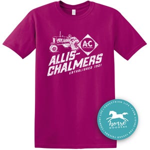 May include: A magenta t-shirt featuring a white graphic of a vintage tractor and the text "ALLIS-CHALMERS ESTABLISHED 1901". A circular logo with a horse graphic is on the bottom right.