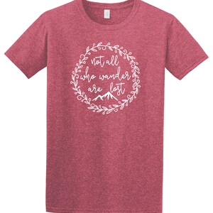以下が含まれることがあります： 白い円形デザインの赤いTシャツ。デザインは、下部に山のイラストが付いた、装飾的なフォントで「not all who wander are lost」というテキストを特徴としています。シャツは半袖とクルーネックです。