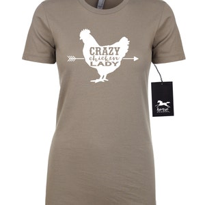 May include: A taupe-colored t-shirt featuring a white graphic of a chicken with the text "CRAZY chickin' LADY" and an arrow. A black tag with a horse logo hangs from the shirt.