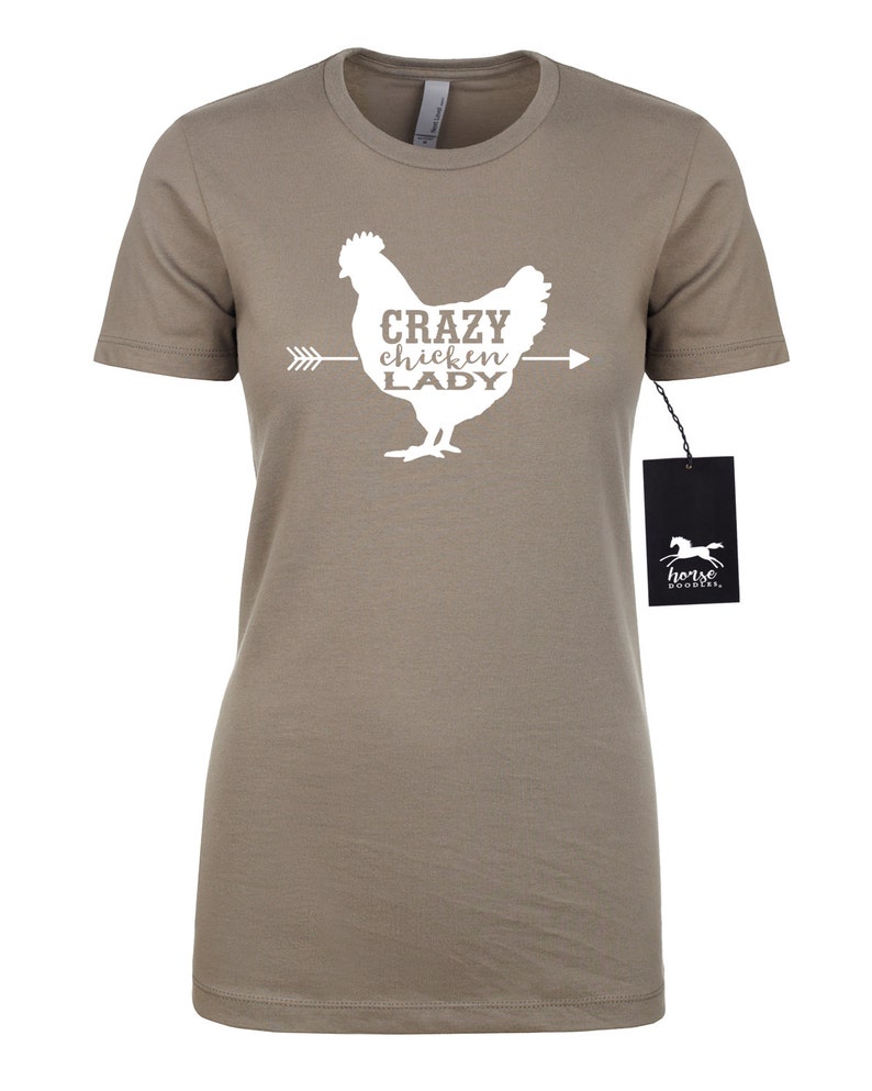 May include: A taupe-colored t-shirt featuring a white graphic of a chicken with the text "CRAZY chickin' LADY" and an arrow. A black tag with a horse logo hangs from the shirt.