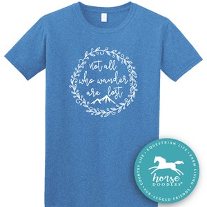 以下が含まれることがあります： 白い花のリースデザインで「not all who wander are lost」というテキストが入った青いTシャツ。シャツには、馬のイラストと「Horse Doodles」という言葉が入った小さな円形のロゴも付いています。