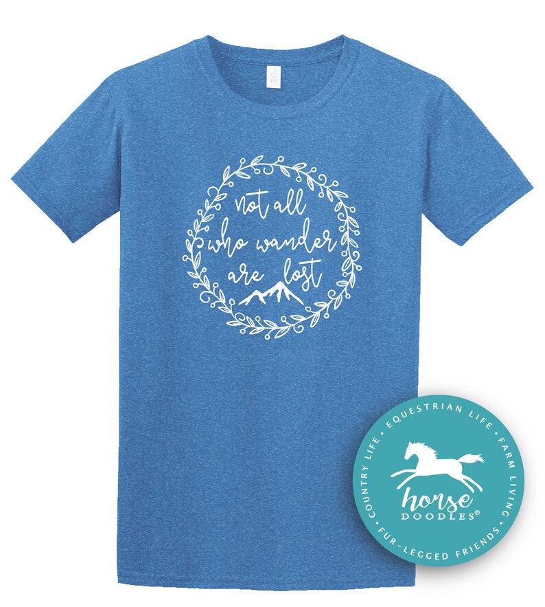 以下が含まれることがあります： 白い花のリースデザインで「not all who wander are lost」というテキストが入った青いTシャツ。シャツには、馬のイラストと「Horse Doodles」という言葉が入った小さな円形のロゴも付いています。